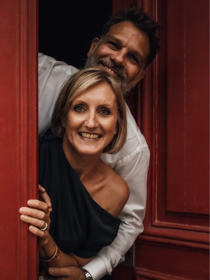 Silvia Vannier, souriante, dans l’embrasure d’une porte rouge, Alexandre derrière elle