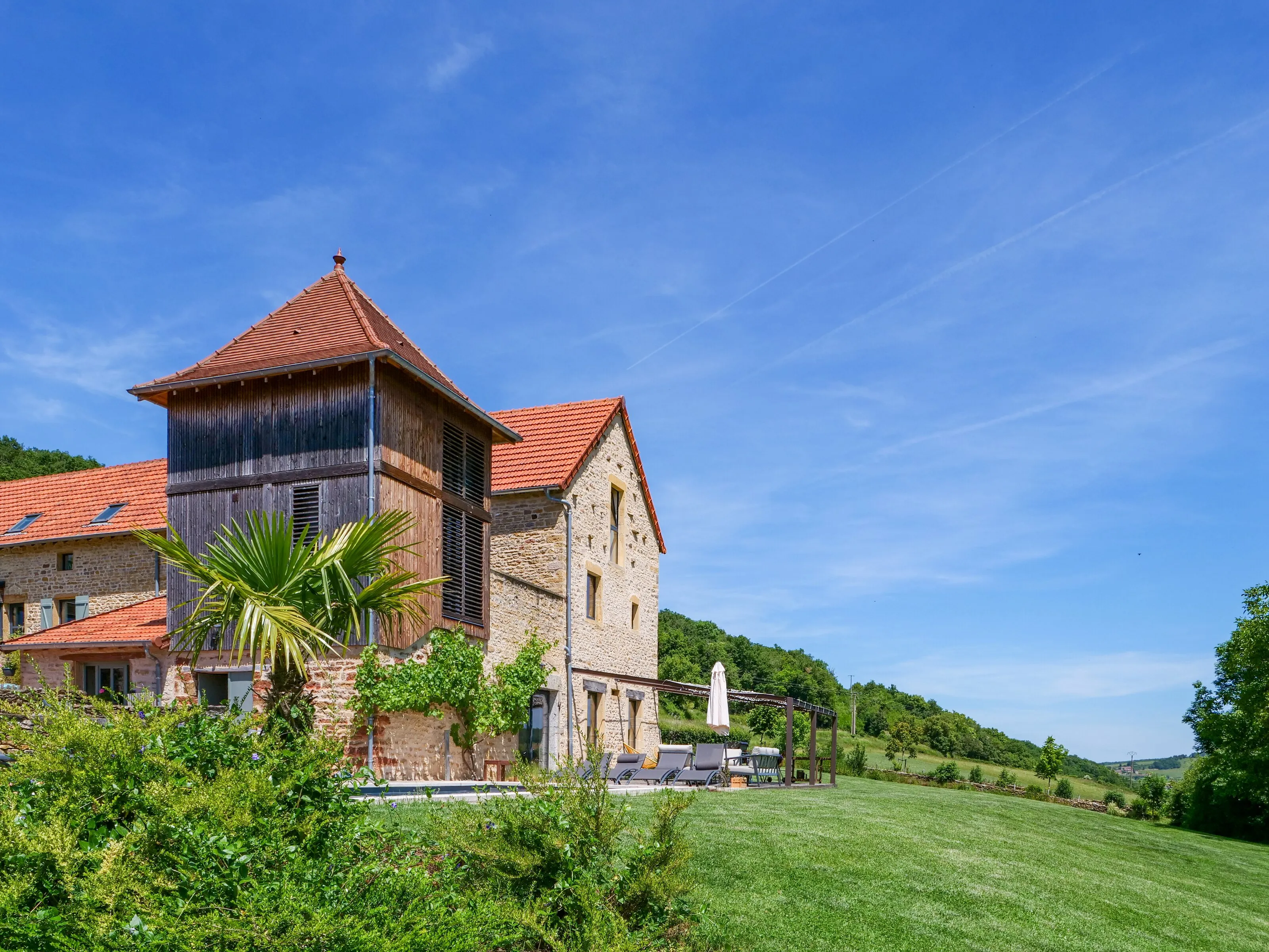 The wooden dovecote and the golden-stone house with a palm tree in front, the lawn and the hills under a wide blue sky